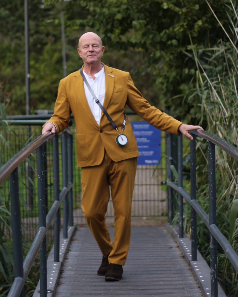 Een man in een okergeel pak staat op een brug tussen het riet, met Het Kompas om zijn nek
